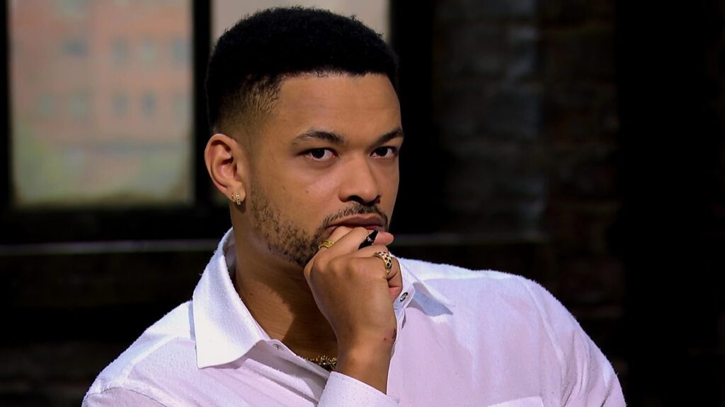 Steven Bartlett on the set of Dragons' Den, wearing a white shirt and gold accessories with a brick wall background.